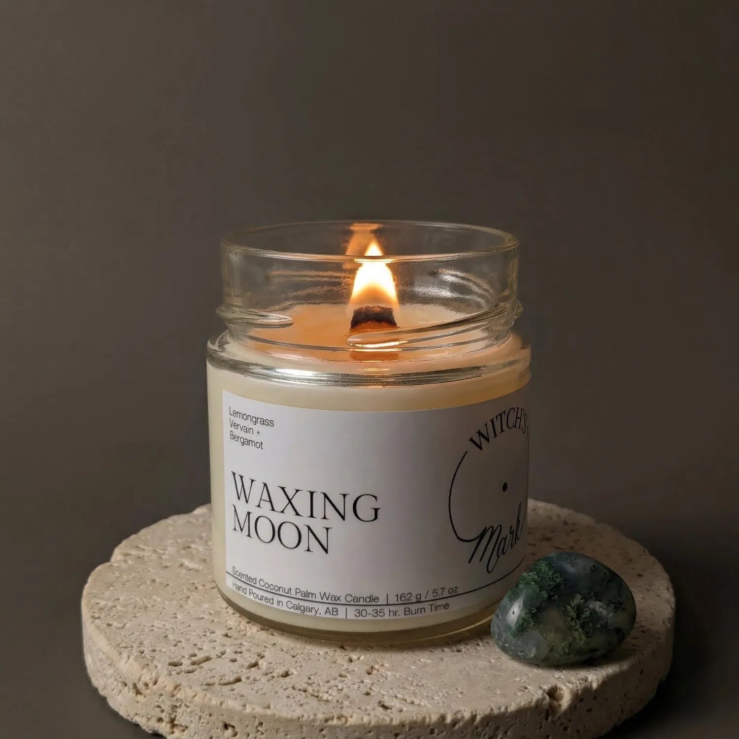 A lit Waxing Moon crystal candle by Witch's Mark resting on a textured stone coaster next to a green moss agate crystal, crafted to help attract wealth and success.