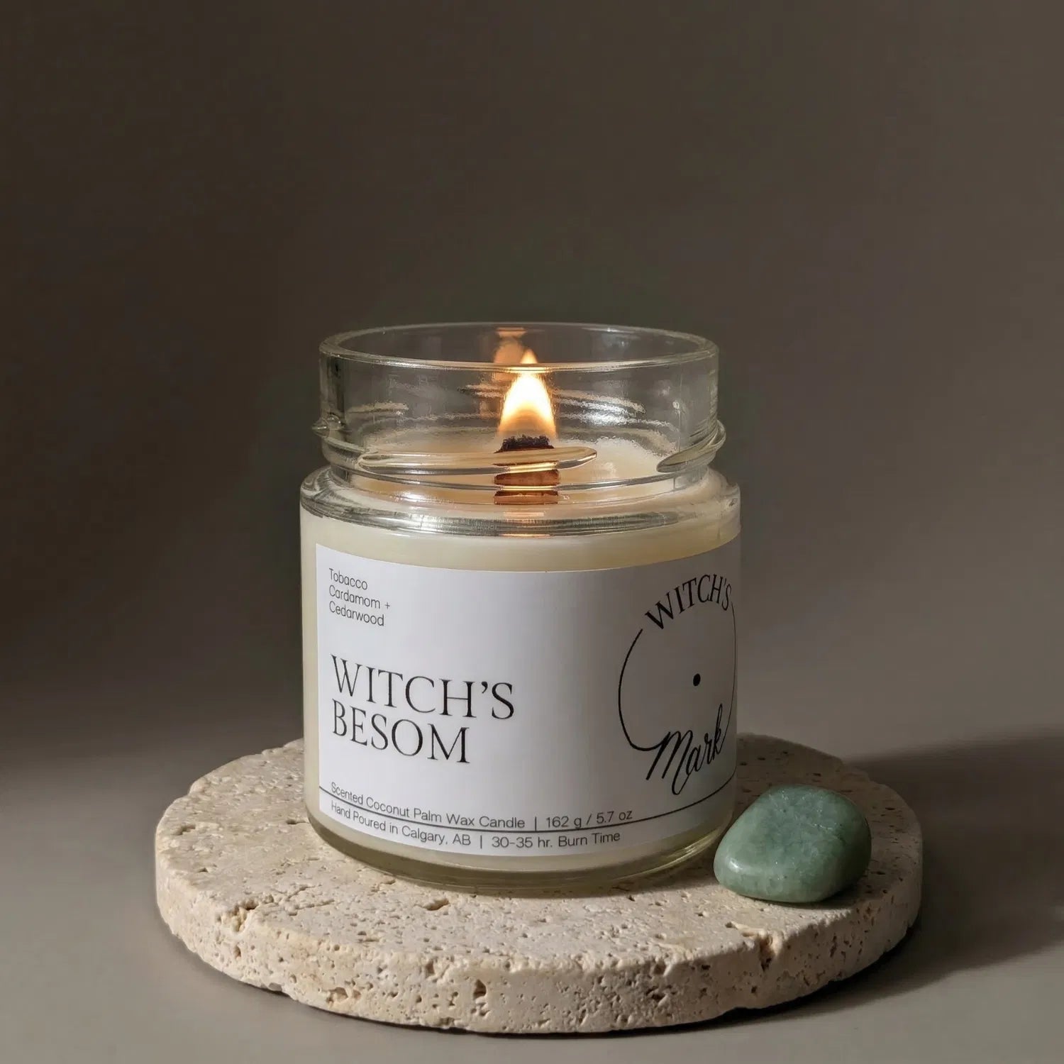 A lit Witch's Besom crystal candle by Witch's Mark resting on a textured stone coaster next to a smooth green jade crystal, crafted to help clear spaces, protect, and attract prosperity.