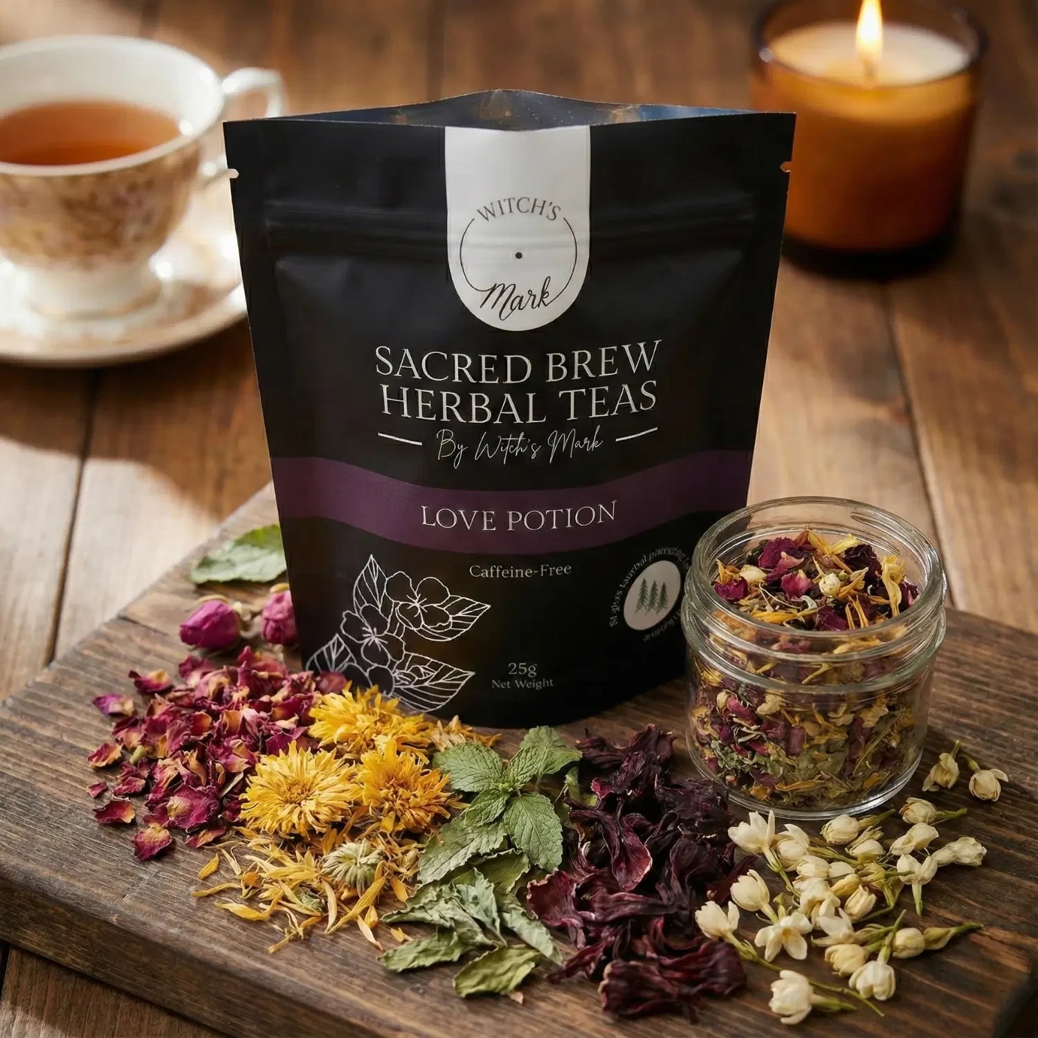 The Sacred Brew Herbal Teas Love Potion pouch by Witch's Mark displayed on a rustic wooden board. This caffeine-free self-care tea blend is surrounded by its raw floral ingredients including vibrant rose petals, golden calendula, damiana, hibiscus, and delicate jasmine blossoms, alongside a warmly lit amber candle and a freshly brewed cup.