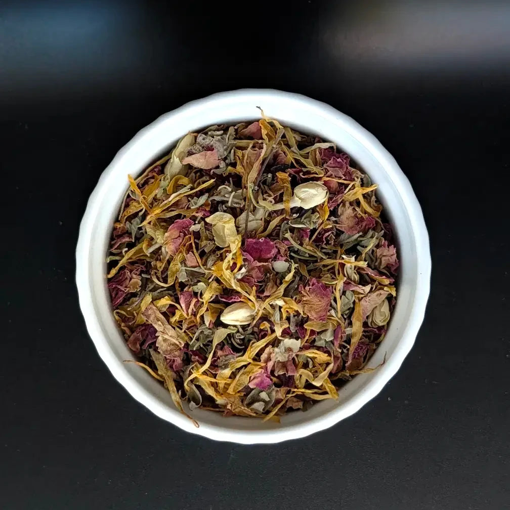 A top-down, close-up view of the Love Potion loose leaf herbal tea blend by Witch's Mark in a small white bowl. The image clearly showcases the high-quality, colorful mix of dried pink rose petals, yellow calendula petals, and green herbal leaves against a dark background.