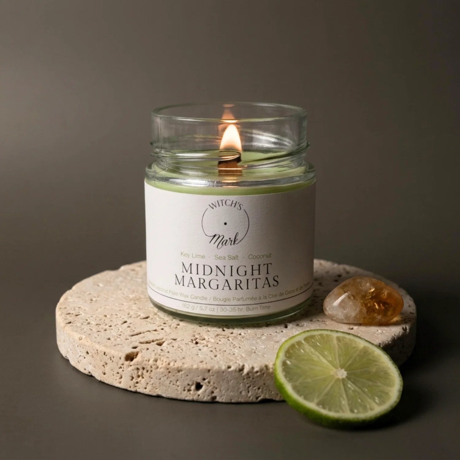 A lit Midnight Margaritas crystal candle by Witch's Mark sitting on a textured stone coaster. A fresh slice of key lime and a radiant citrine crystal rest beside it against a dark, moody background.