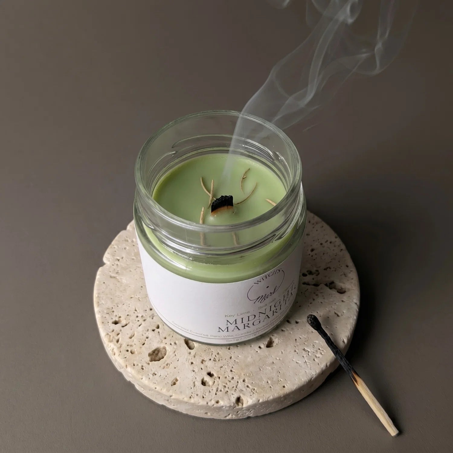 A freshly extinguished Midnight Margaritas candle emitting a trail of smoke from its wooden wick, resting on a stone coaster with a burnt match nearby.