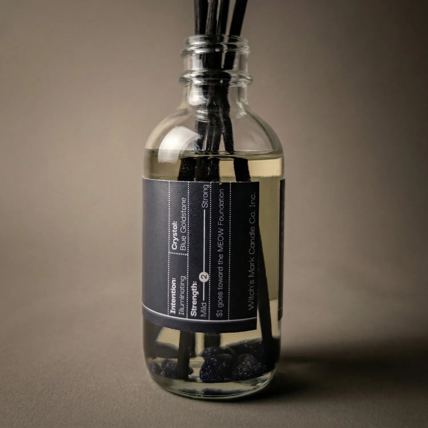 A warmly lit close-up of the Midnight Séance spell jar reed diffuser, revealing the shimmering blue goldstone crystals submerged at the bottom of the clear botanical oil, crafted to provide profound cosmic insight and illuminate the unknown.