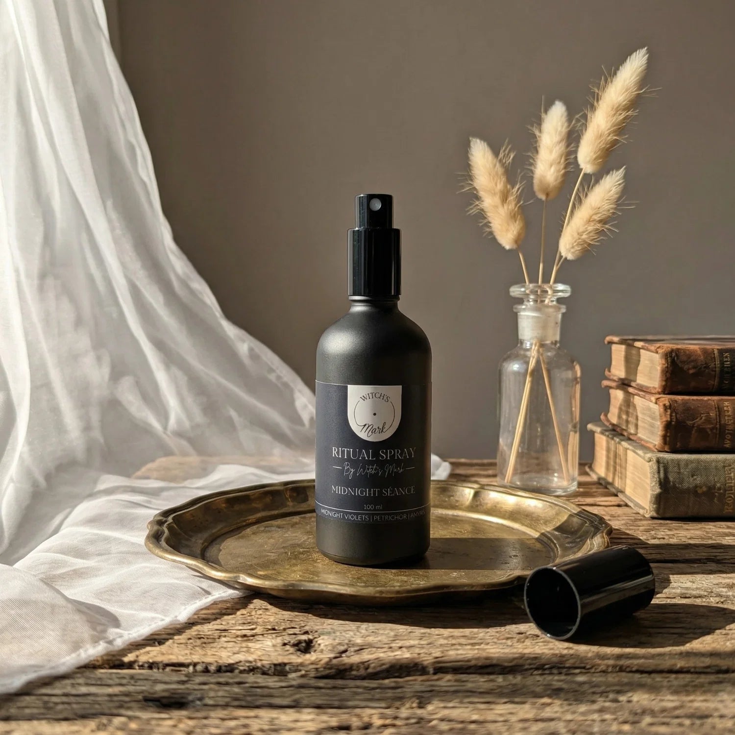 The matte black bottle of Midnight Séance ritual spray with its dark blue label elegantly displayed on a vintage brass tray atop a rustic wooden table. This sacred elixir is perfect for misting over your altar or tarot deck to clear mental static and invoke whispered mystery.