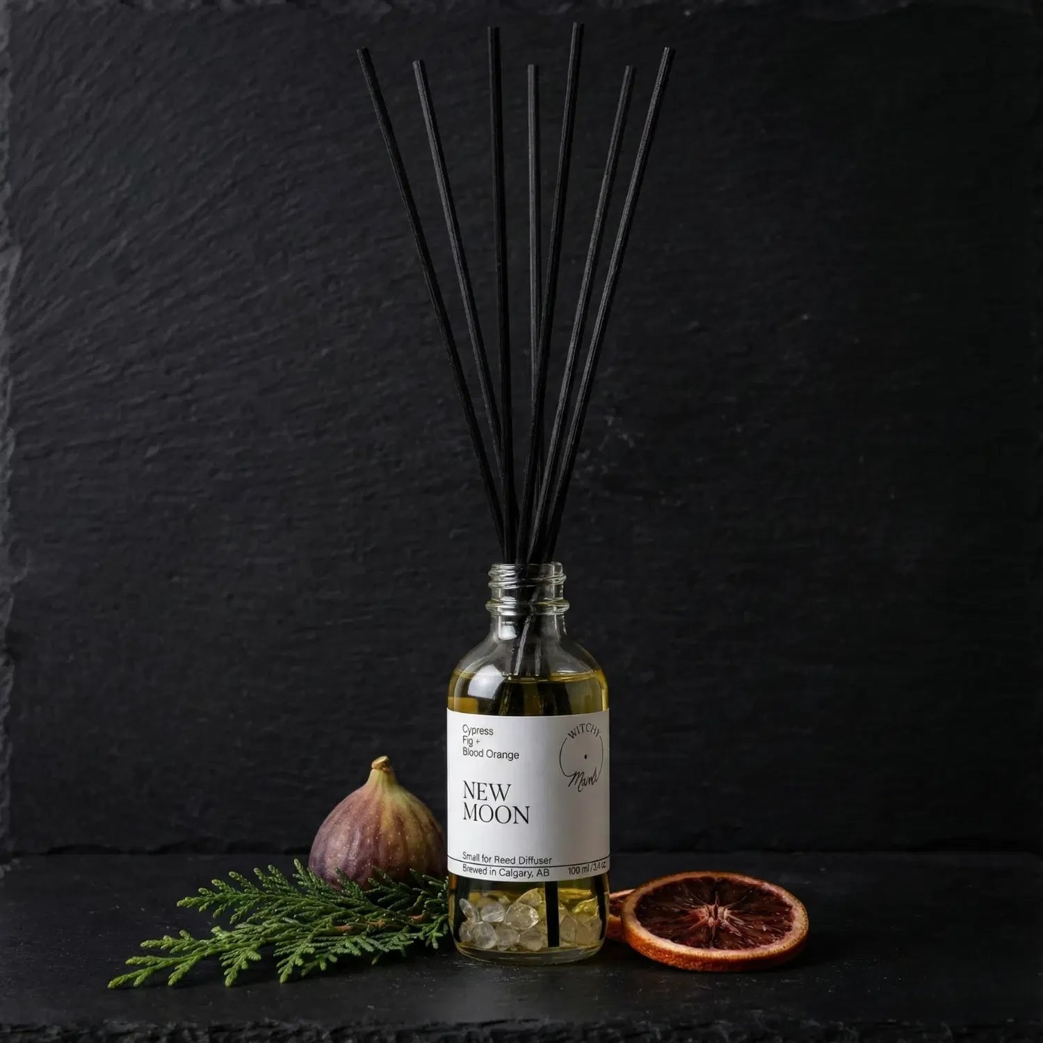 The New Moon spell jar reed diffuser by Witch's Mark showcased against a dark slate background, surrounded by its core botanical ingredients: a fresh purple fig, a sprig of green cypress, and a slice of dried blood orange.