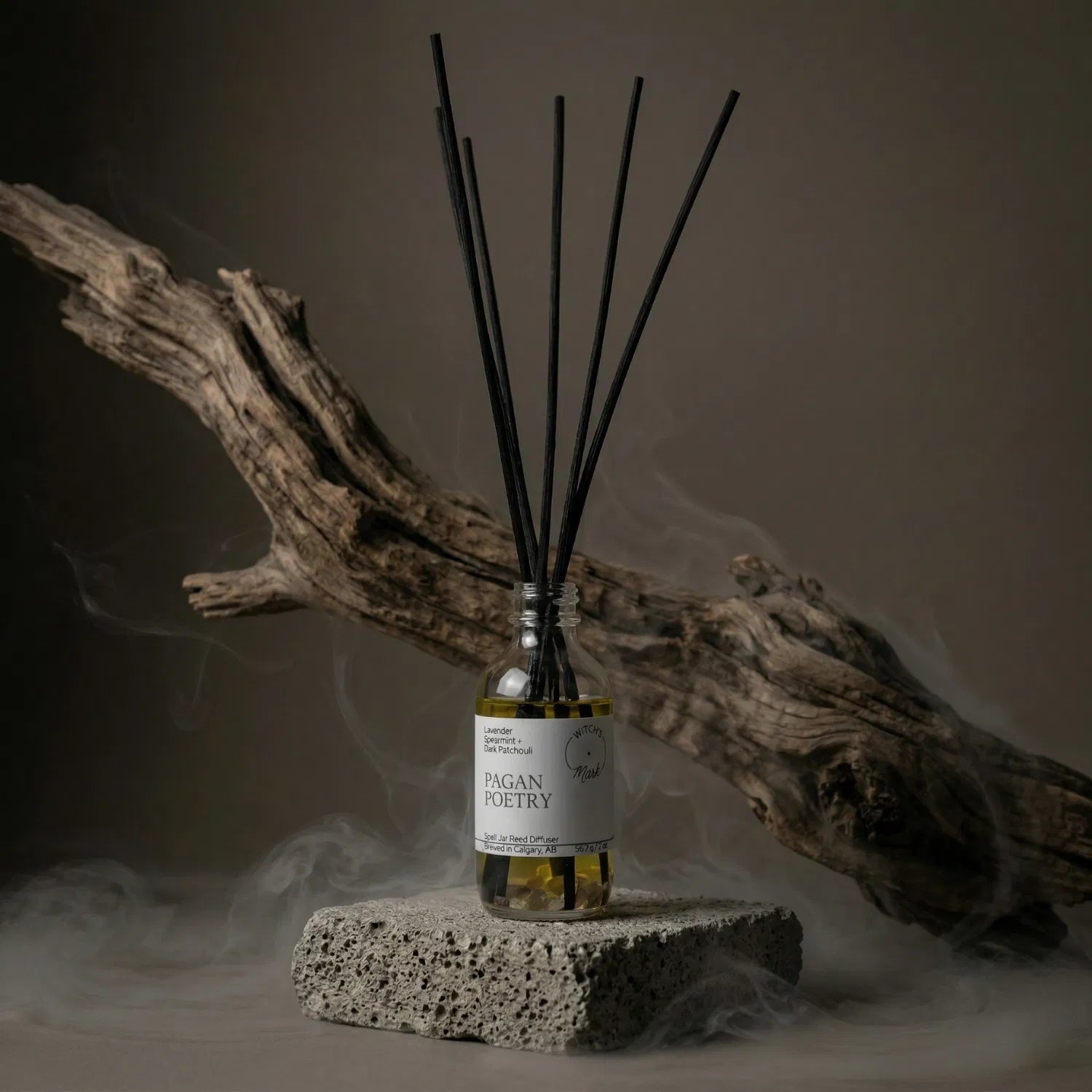 The Pagan Poetry apothecary spell jar reed diffuser by Witch's Mark sitting on a stone base, enveloped in atmospheric mist with natural driftwood in the background, perfectly setting the stage for solitary reflection and a grounded sanctuary.