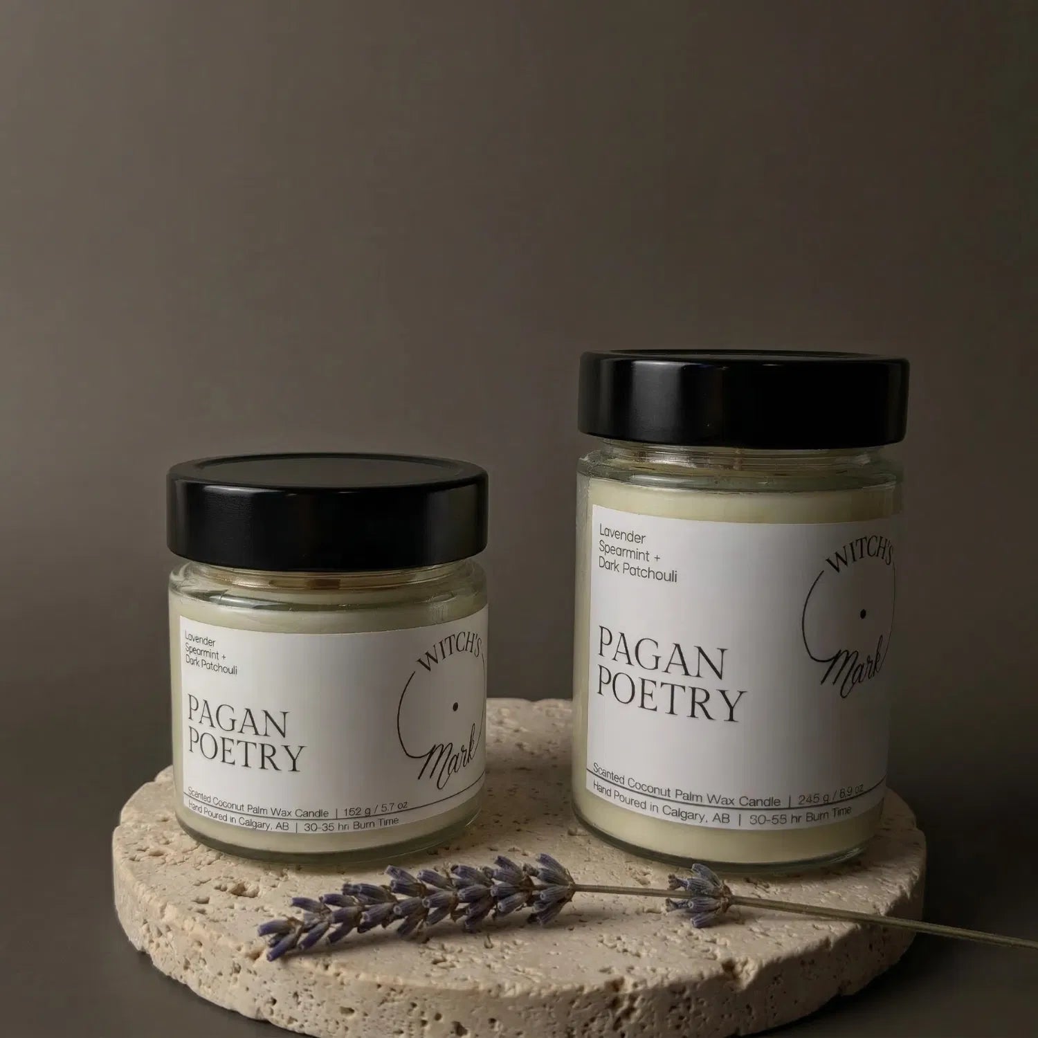 Two sizes of the Witch's Mark Pagan Poetry coconut wax candle, 5.7 oz and 8.6 oz, displayed on a round stone base and garnished with a sprig of dried lavender.