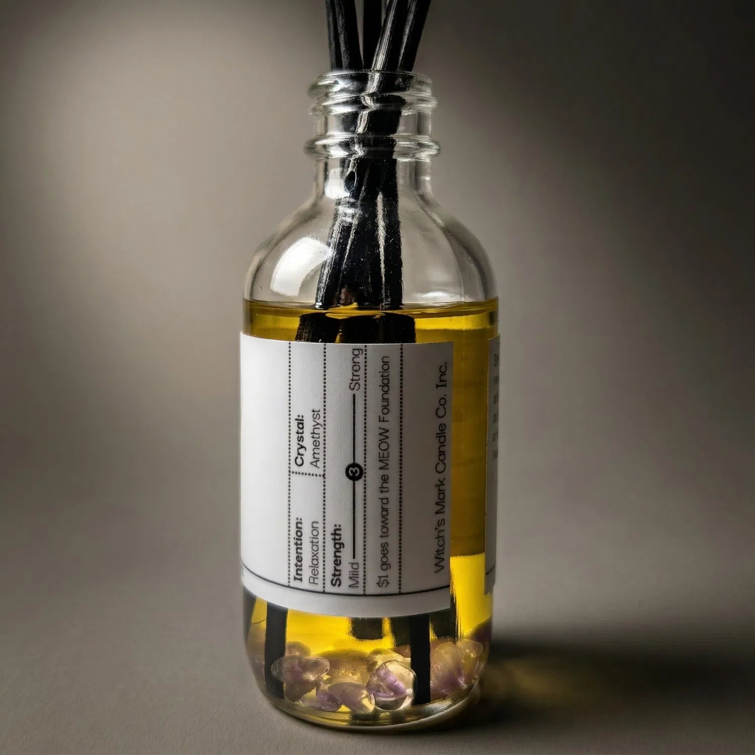 A warmly lit close-up of the Pagan Poetry spell jar reed diffuser, revealing the soothing amethyst crystals submerged at the bottom of the golden botanical oil, crafted to ease an anxious spirit and deepen intuition.