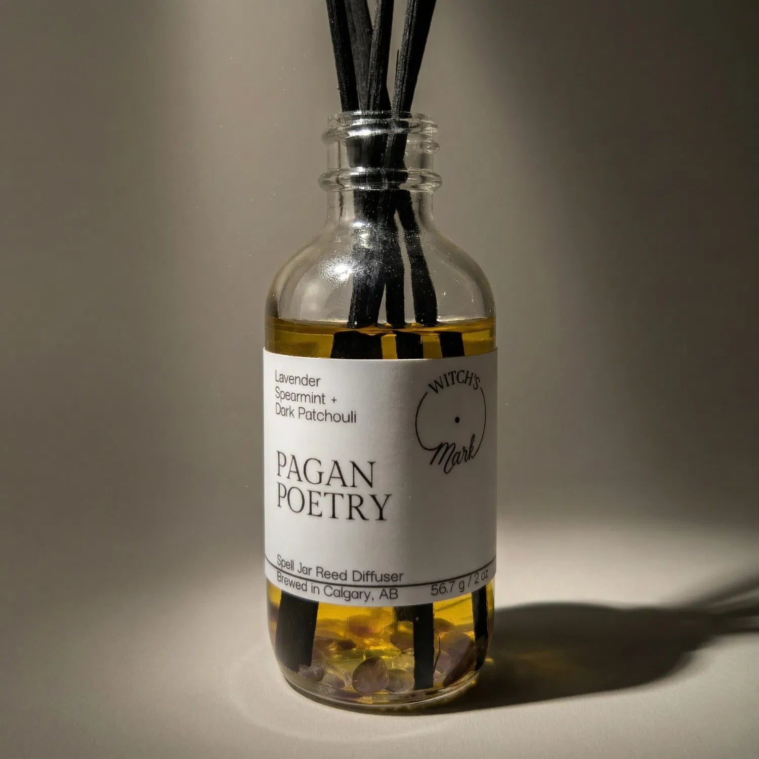 A dramatic spotlight close-up of the Pagan Poetry spell jar reed diffuser, highlighting the front label which lists its magical, tranquility-inducing scent profile of calming lavender, crisp spearmint, and dark patchouli.