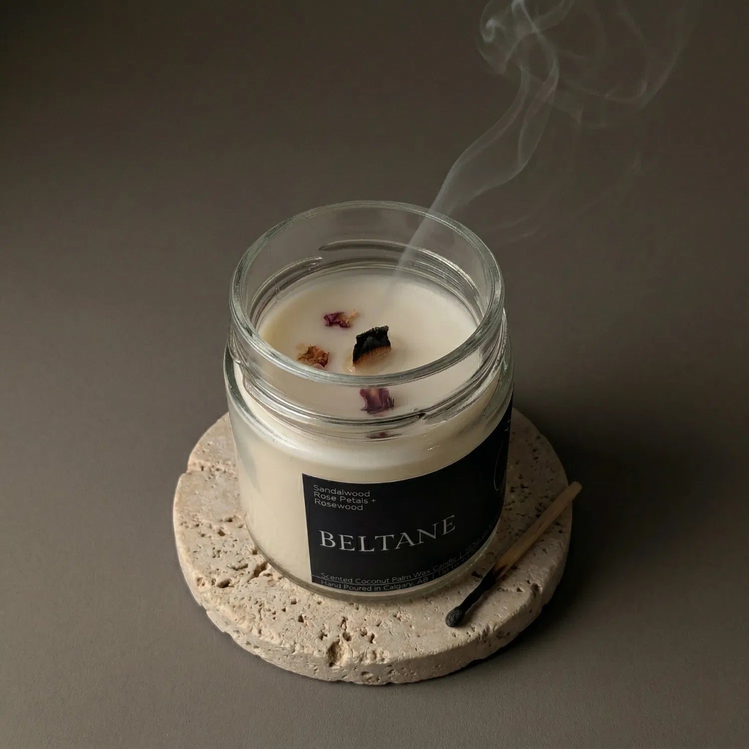 A freshly extinguished Beltane candle resting on a stone coaster, emitting a delicate trail of calming smoke from its wooden wick, with a burnt match resting nearby.