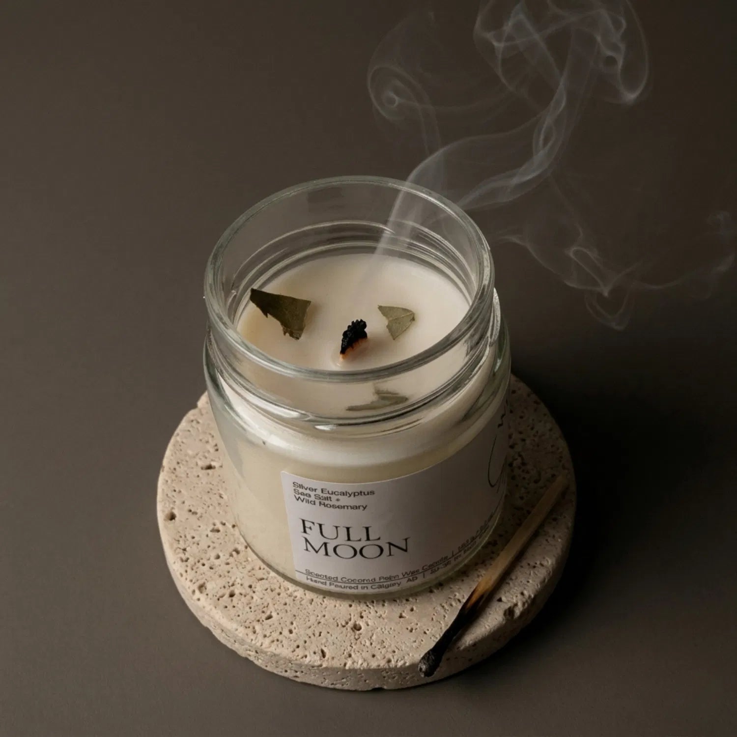 A freshly extinguished Full Moon candle resting on a stone coaster, emitting a delicate trail of purifying smoke from its wooden wick, with a burnt match resting nearby.