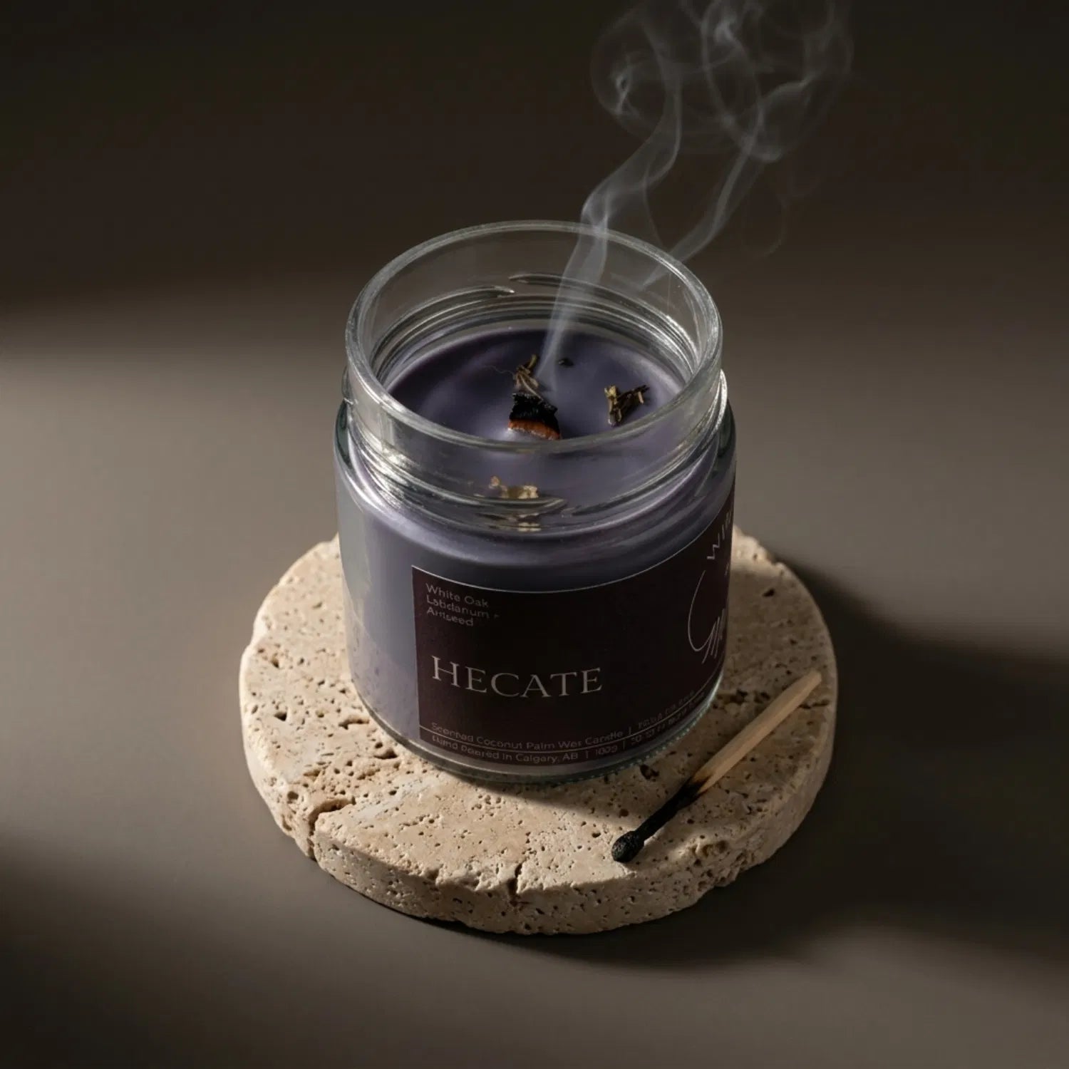 A freshly extinguished Hecate candle resting on a stone coaster, emitting a delicate trail of smoke from its wooden wick after a psychic enhancement ritual. The purple wax is lightly adorned with mugwort, with a burnt match resting nearby.