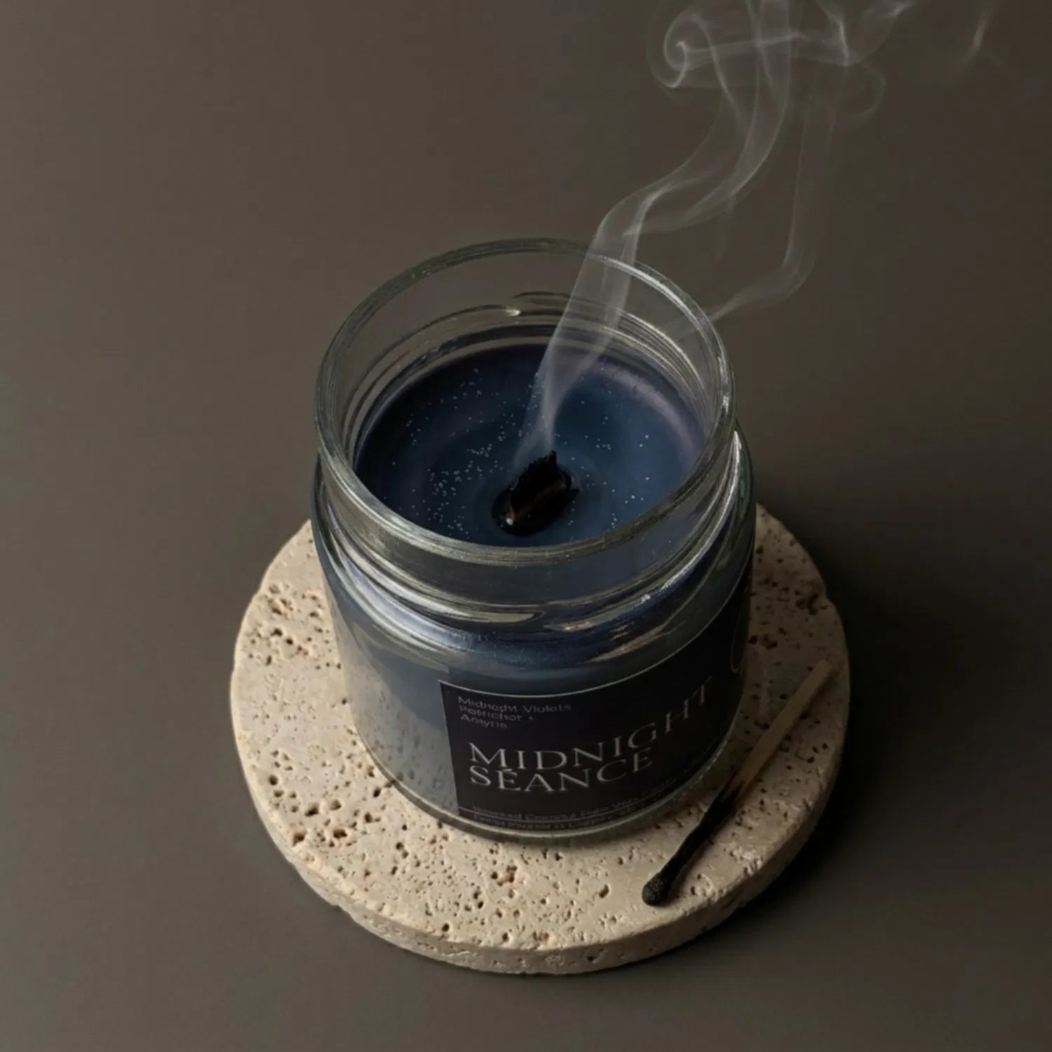 A freshly extinguished Midnight Séance candle resting on a stone coaster, emitting a delicate trail of calming smoke from its wooden wick after a spiritual practice, with a burnt match resting nearby.