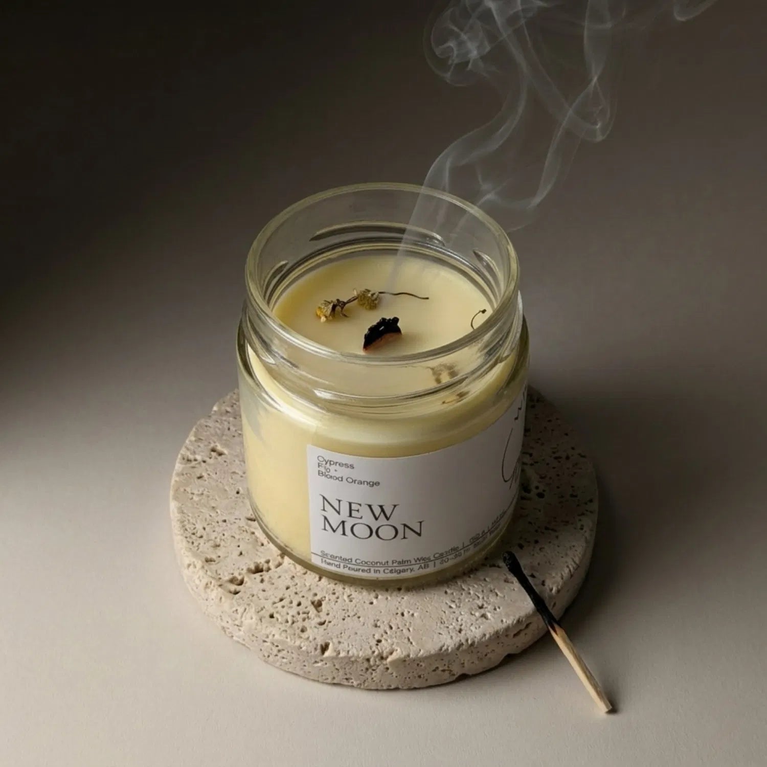 A freshly extinguished New Moon candle resting on a stone coaster, emitting a delicate trail of smoke from its wooden wick, with a burnt match resting nearby.