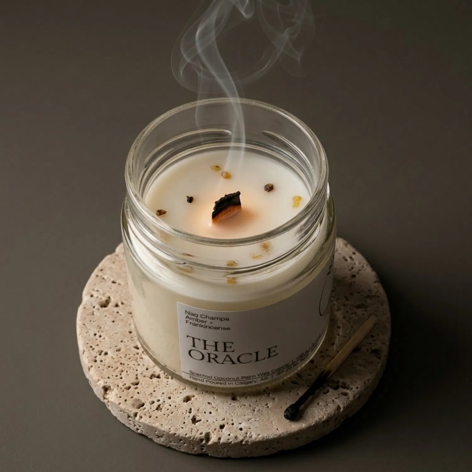 A freshly extinguished Oracle candle resting on a stone coaster, emitting a trail of calming smoke from its wooden wick after a meditation session, with a burnt match resting nearby.