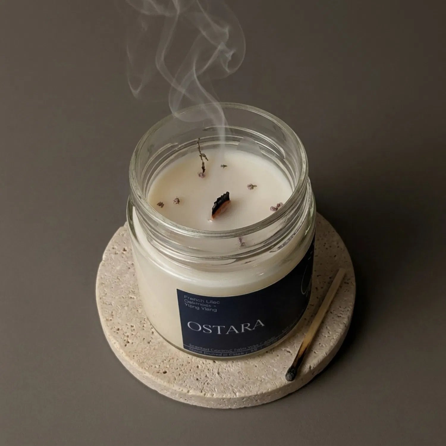 A freshly extinguished Ostara candle resting on a stone coaster, emitting a delicate trail of relaxing smoke from its wooden wick, with a burnt match resting nearby.