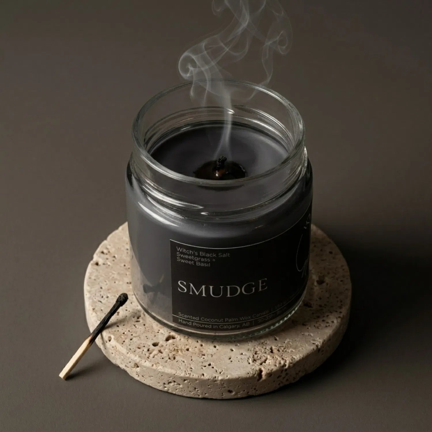A freshly extinguished Smudge candle emitting a trail of purifying smoke from its wooden wick, resting on a stone coaster with a burnt match nearby.