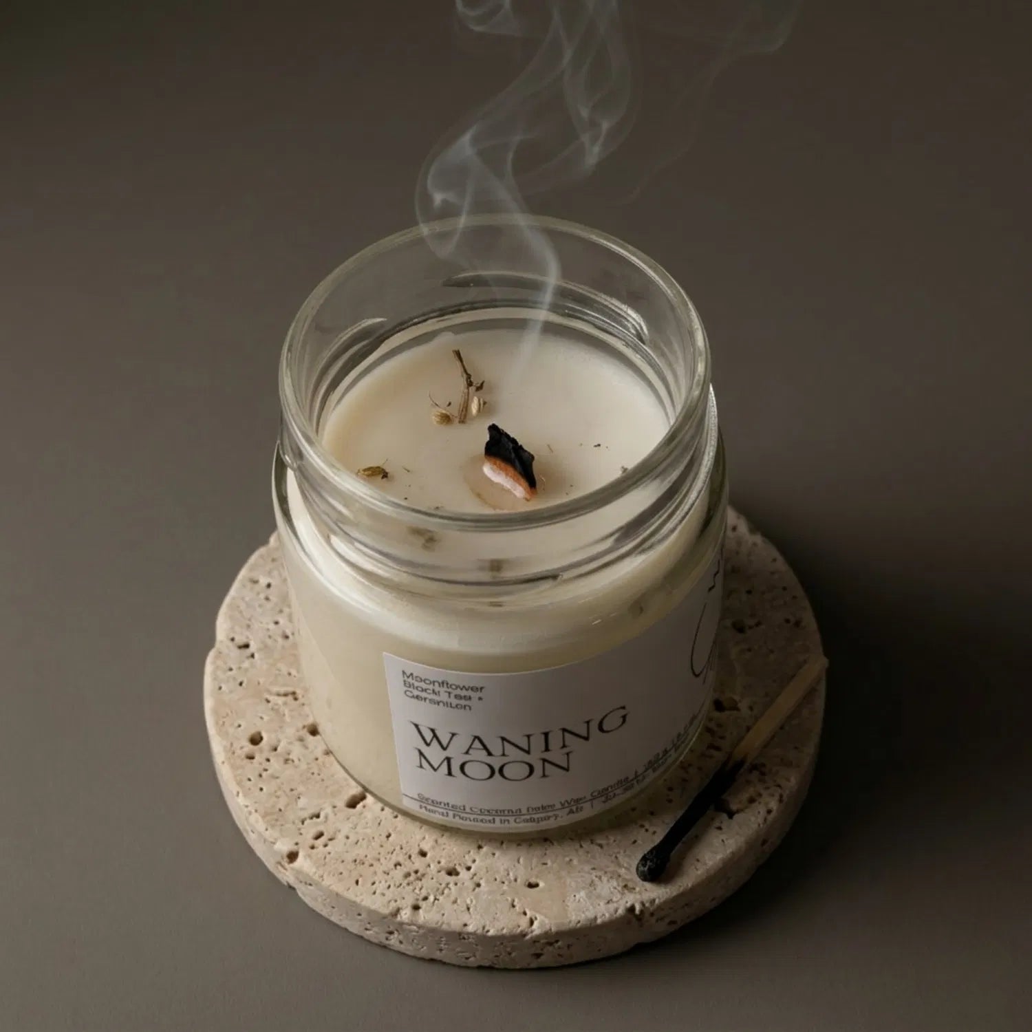 A freshly extinguished Waning Moon candle resting on a stone coaster, emitting a delicate trail of calming smoke from its wooden wick, with a burnt match resting nearby.