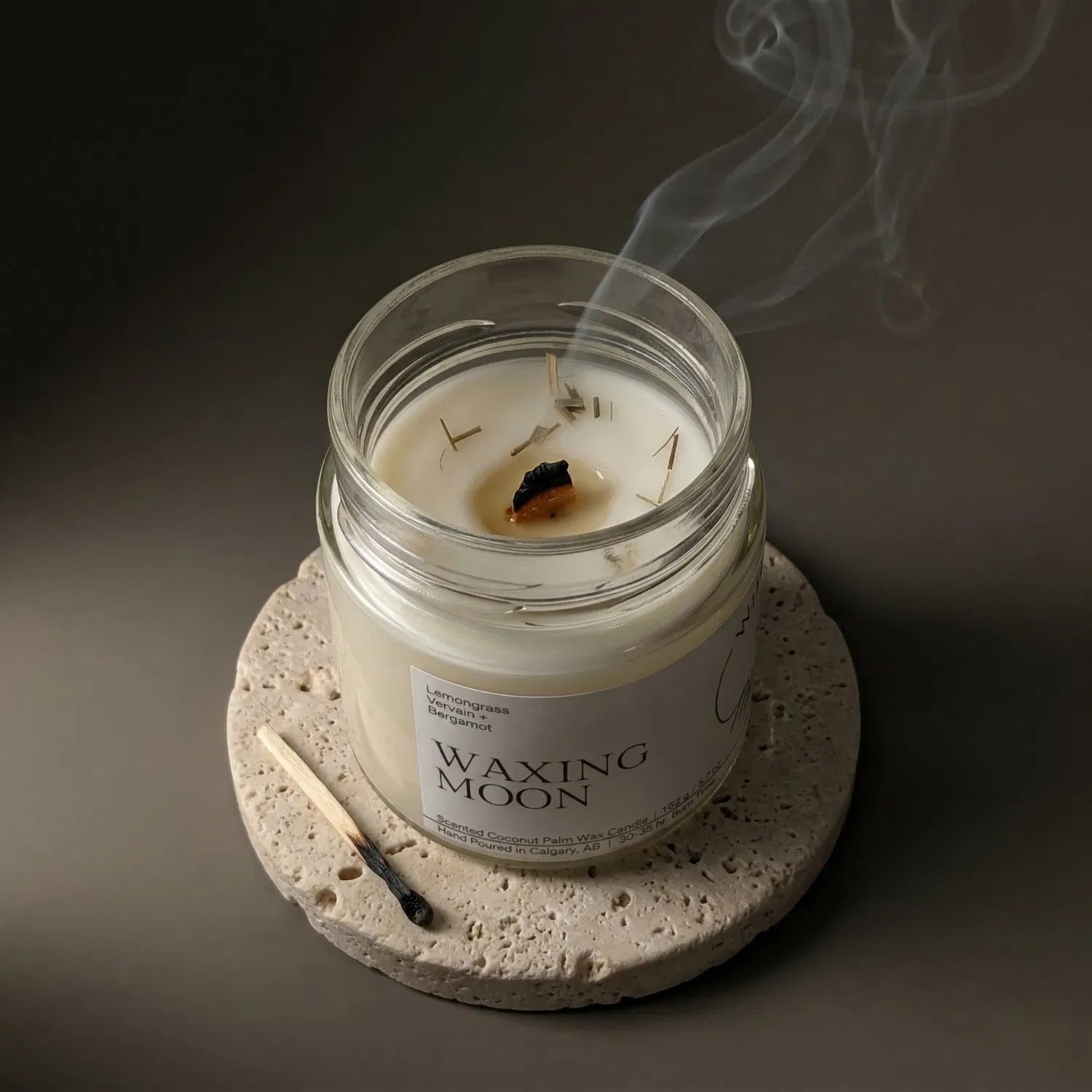 A freshly extinguished Waxing Moon candle resting on a stone coaster, emitting a delicate trail of smoke from its wooden wick, with a burnt match resting nearby.