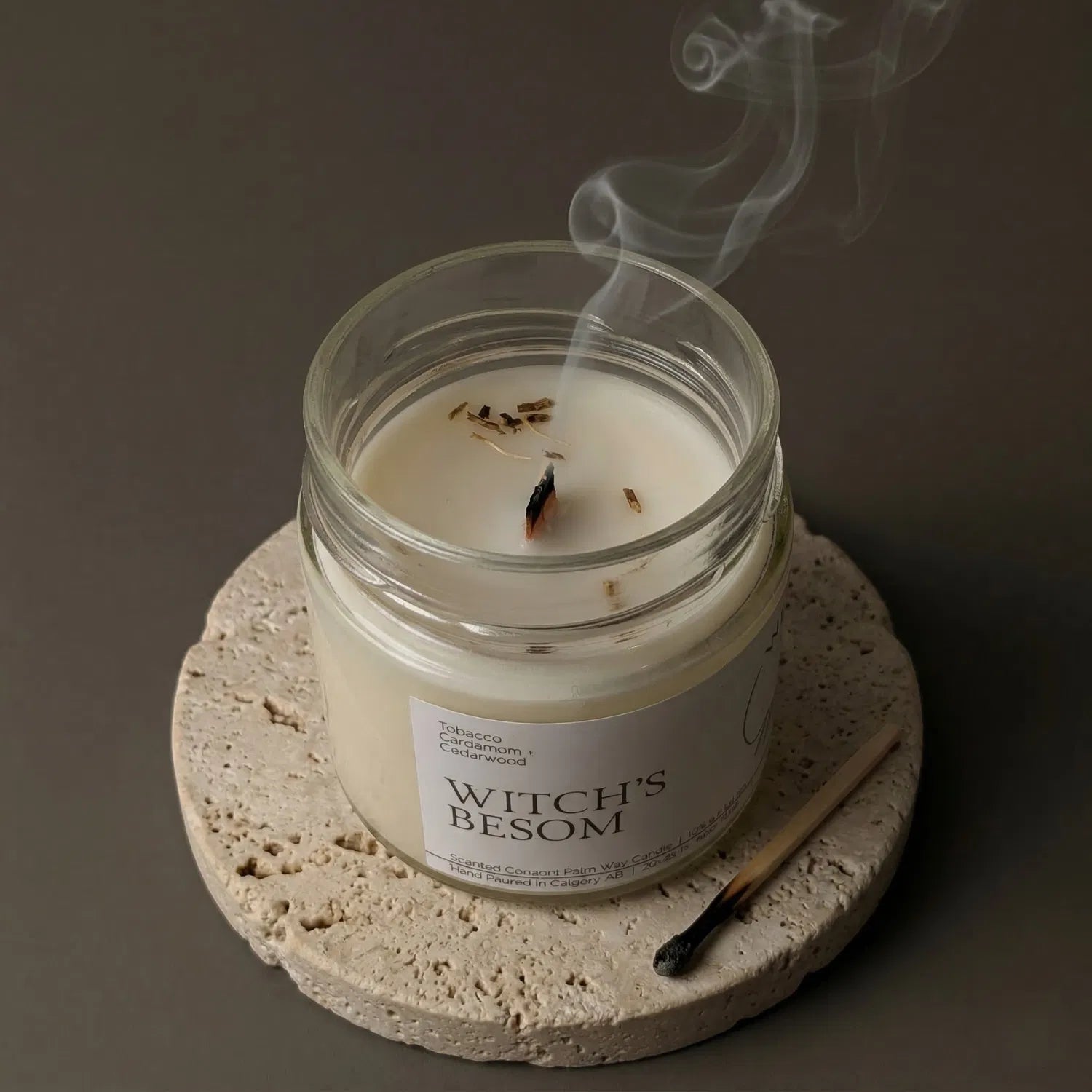 A freshly extinguished Witch's Besom candle resting on a stone coaster, emitting a delicate trail of purifying smoke from its wooden wick, with a burnt match resting nearby.