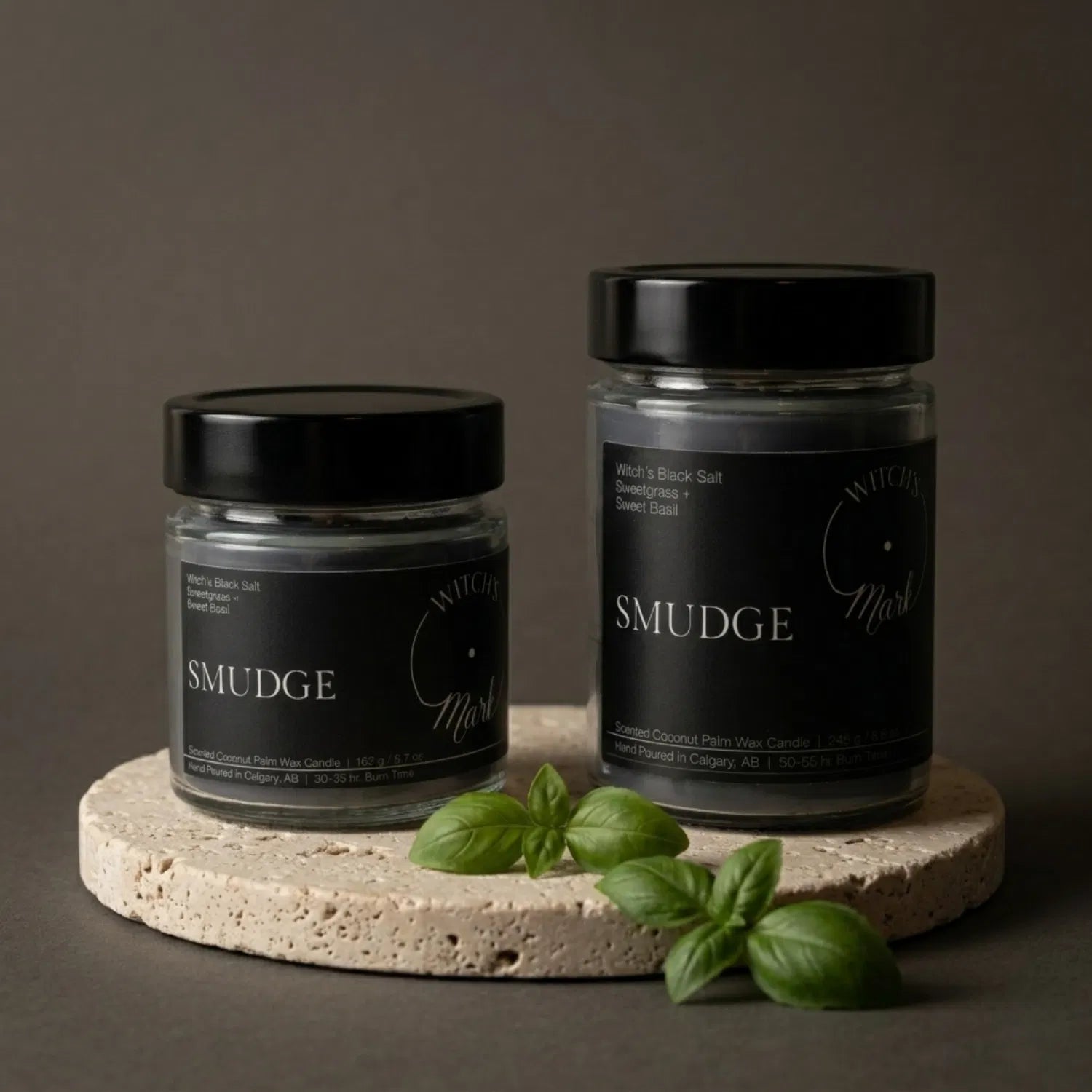 Two sizes of the Witch's Mark Smudge coconut wax candle, 5.7 oz and 8.6 oz, displayed on a round stone base and garnished with fresh sweet basil leaves.