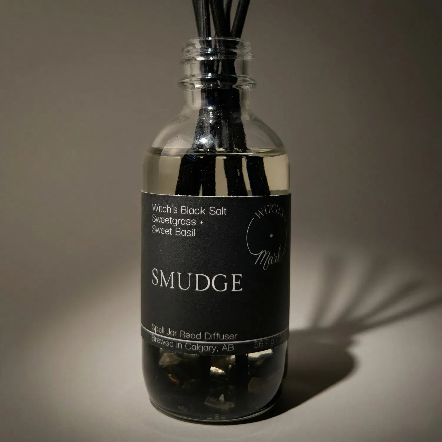 A dramatic spotlight close-up of the Smudge spell jar reed diffuser, highlighting the dark front label which lists its magical, purifying scent profile of witch's black salt, sacred sweetgrass, and sweet basil.