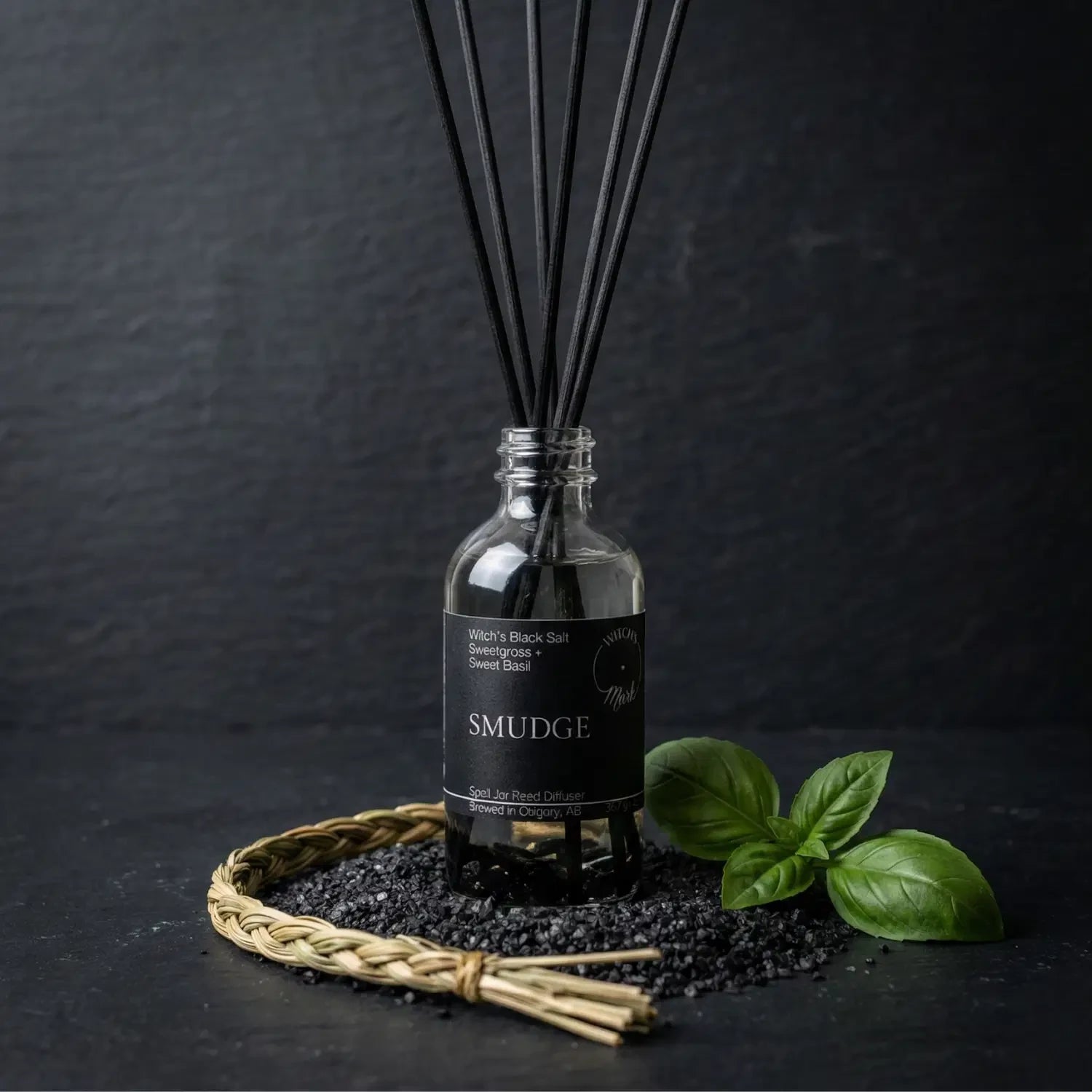 The Smudge spell jar reed diffuser by Witch's Mark showcased against a dark slate background, surrounded by its core protective botanical ingredients: a braid of sacred sweetgrass, a scattering of witch's black salt, and fresh green sweet basil leaves.