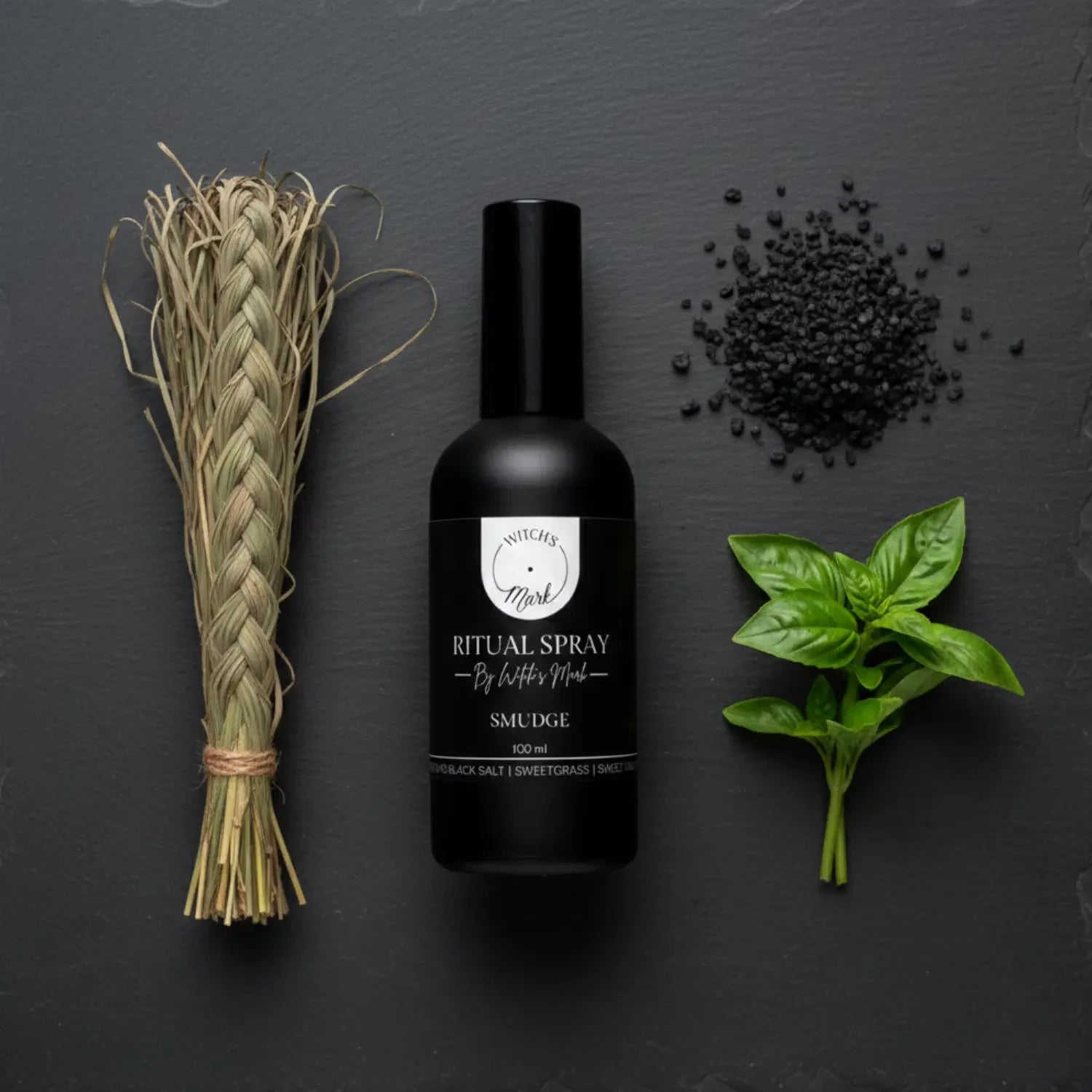 A top-down flat lay highlighting the potent ingredients of the Smudge ritual mist by Witch's Mark, featuring the matte black bottle surrounded by a braid of sacred sweetgrass, a scattering of witch's black salt, and fresh sweet basil leaves.