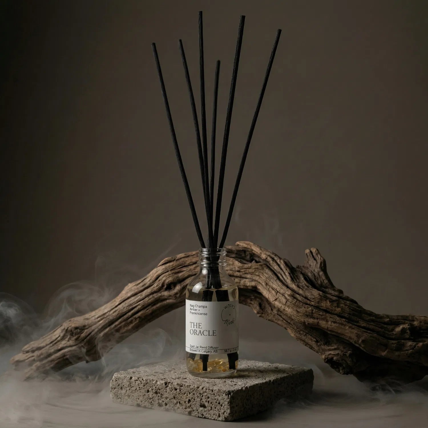 The Oracle apothecary spell jar reed diffuser by Witch's Mark sitting on a stone base, enveloped in atmospheric mist with natural driftwood in the background, perfectly setting the stage for tarot readings and sharpening mental focus.