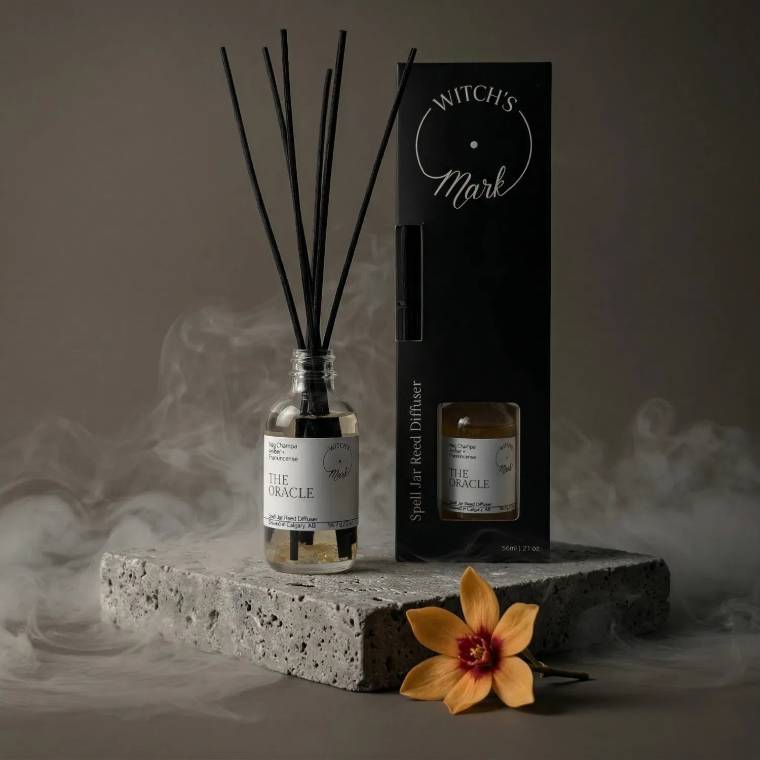 The Oracle spell jar reed diffuser by Witch's Mark displayed next to its sleek black packaging box. The apothecary glass vessel with black reeds rests on a textured stone block surrounded by mystical mist and a fresh yellow champaca flower, perfect for setting a divination space.