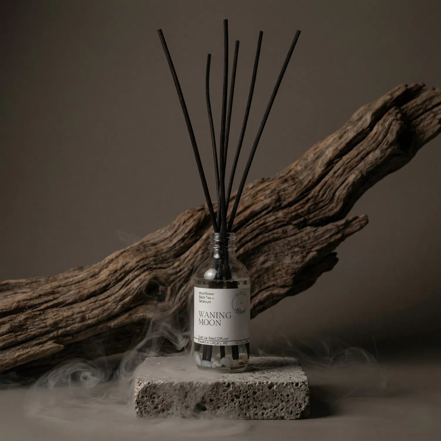 The Waning Moon apothecary spell jar reed diffuser by Witch's Mark sitting on a stone base, enveloped in atmospheric mist with natural driftwood in the background, perfectly setting the stage for deep spiritual release and journeying through shadow phases.