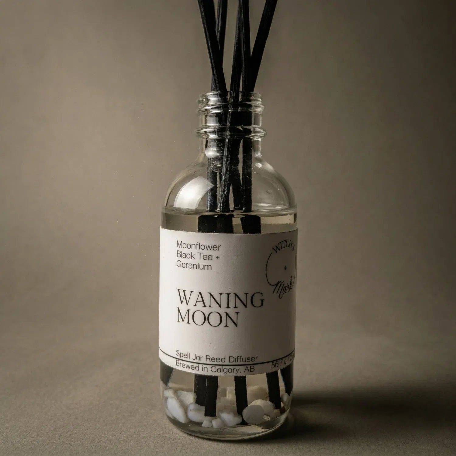 A dramatic spotlight close-up of the Waning Moon spell jar reed diffuser, highlighting the front label which lists its magical, restorative scent profile of night-blooming moonflower, black tea, and geranium.