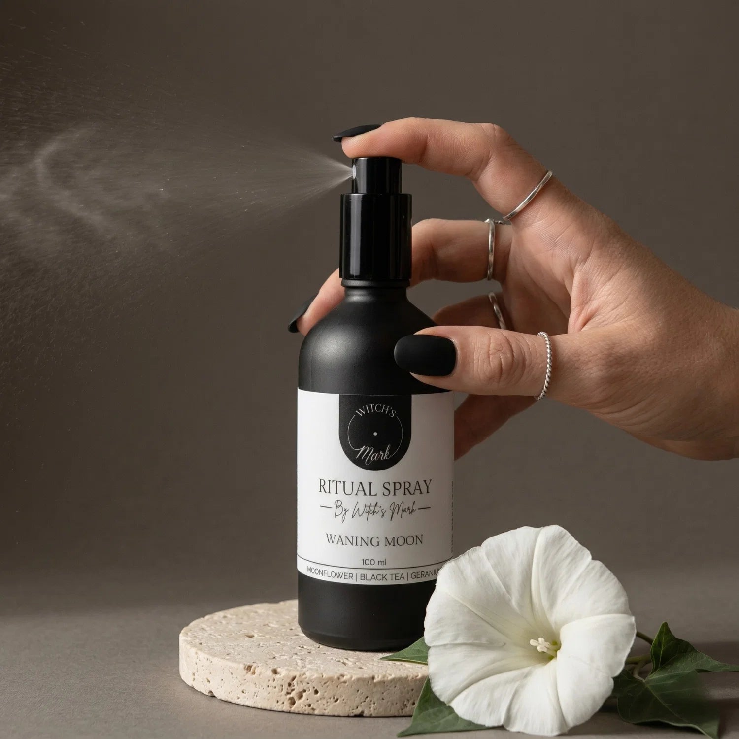 A hand with black nail polish spritzing the Witch's Mark Waning Moon ritual spray over a fresh white moonflower. This calming aura mist is infused with polychrome jasper charged moon water and essential oils to support deep emotional unburdening and necessary release.
