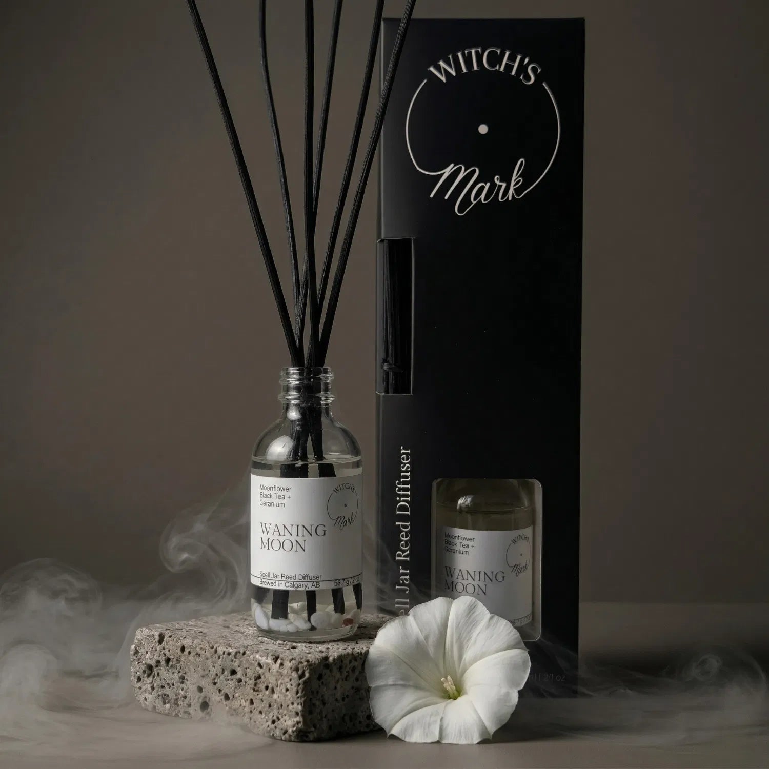 The Waning Moon spell jar reed diffuser by Witch's Mark displayed next to its sleek black packaging box. The apothecary glass vessel with black reeds rests on a textured stone block surrounded by mystical mist and a delicate white moonflower, perfect for rituals of release.
