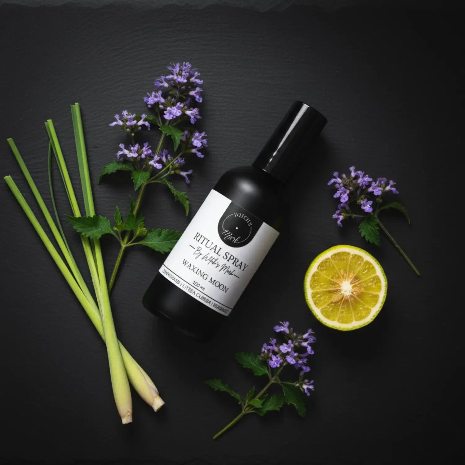 A top-down flat lay highlighting the uplifting ingredients of the Waxing Moon ritual mist by Witch's Mark, featuring the black apothecary bottle surrounded by fresh lemongrass stalks, a slice of bright bergamot, and delicate purple botanicals against a dark slate background.