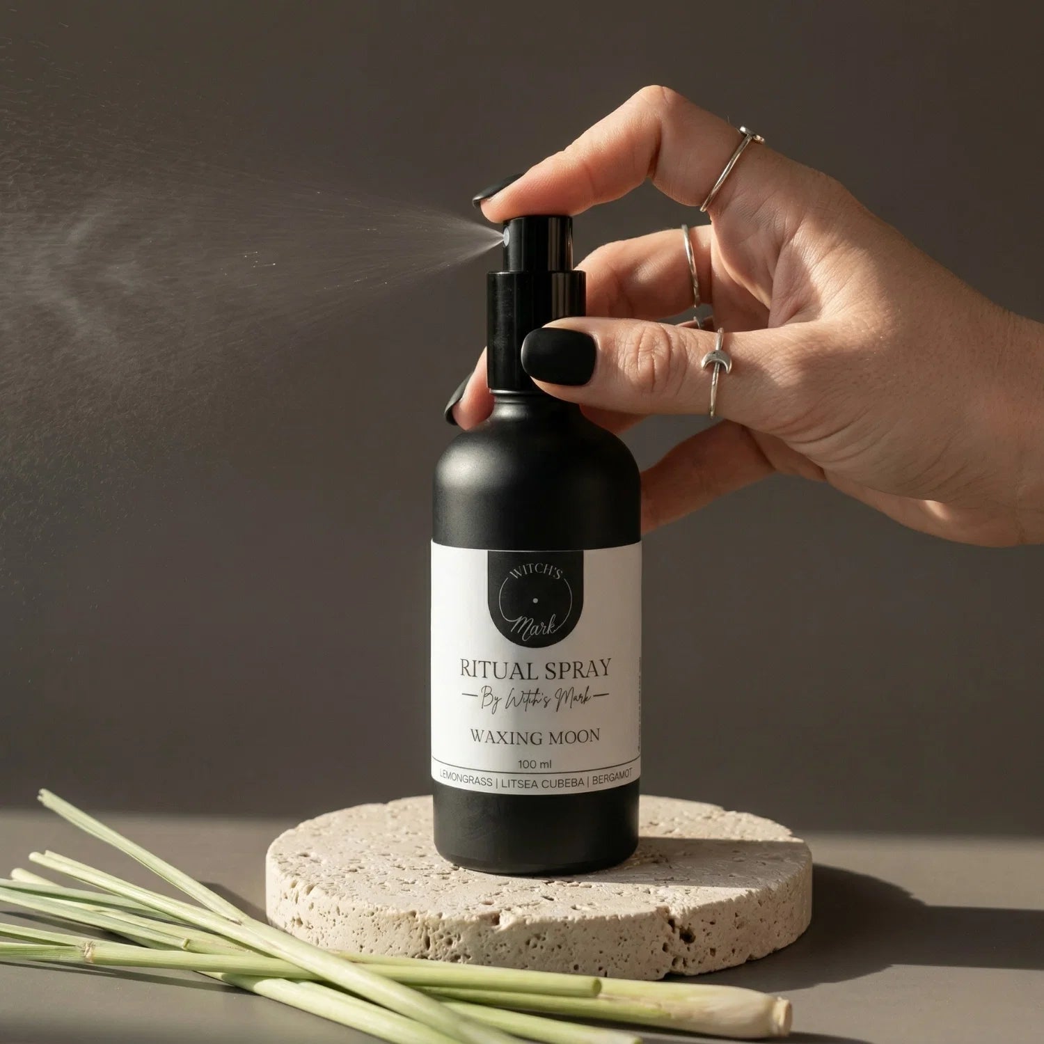 A hand with black nail polish spritzing the Witch's Mark Waxing Moon ritual spray over fresh stalks of lemongrass. This uplifting aura mist is infused with moss agate moon water and pure essential oils to fuel motivation and amplify manifestation magick.