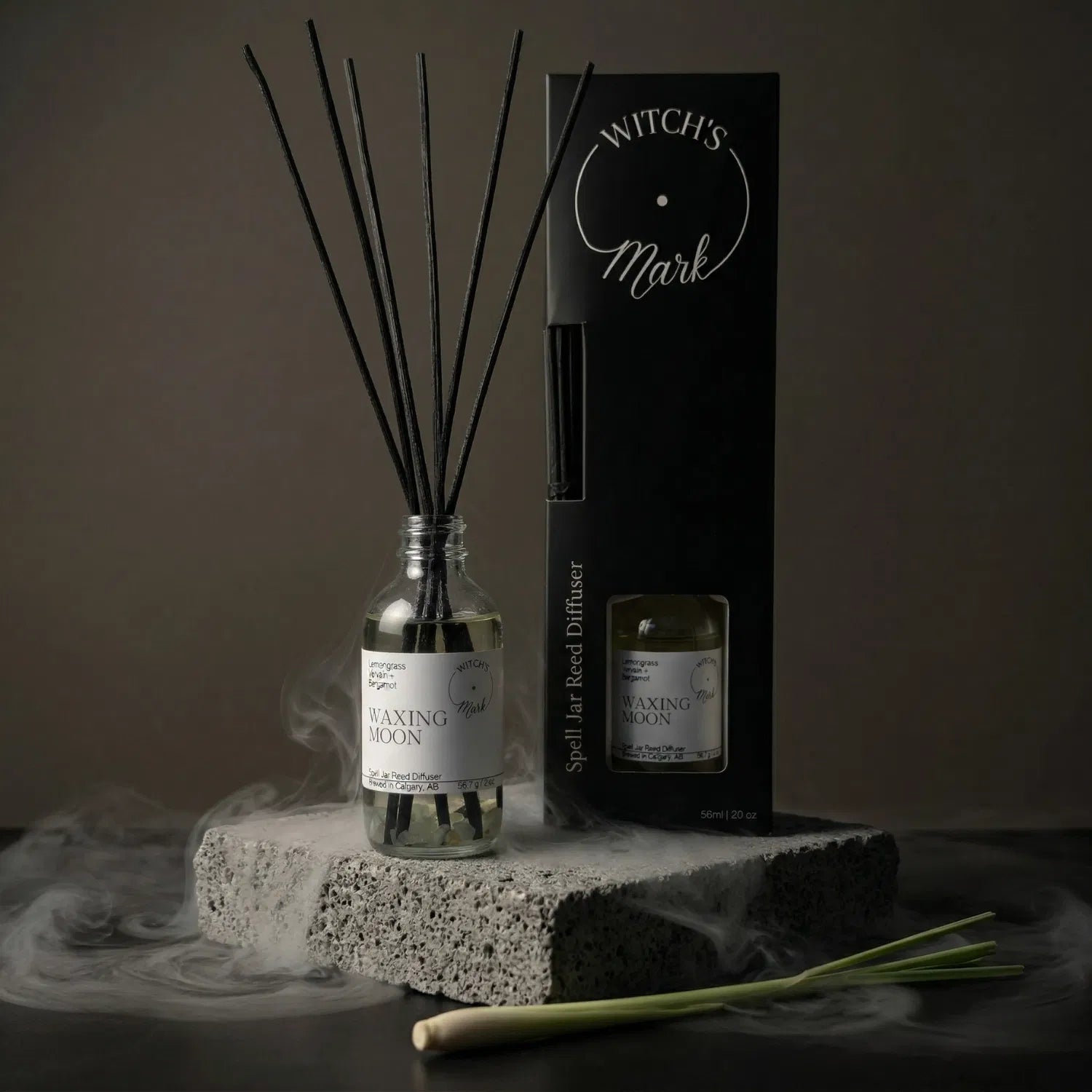 The Waxing Moon spell jar reed diffuser by Witch's Mark displayed next to its sleek black packaging box. The apothecary glass vessel with black reeds rests on a textured stone block surrounded by mystical mist and a fresh stalk of grounding lemongrass.