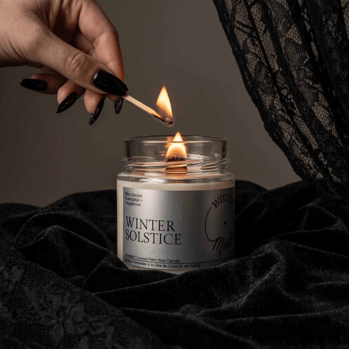A hand with black manicured nails lights a candle in a clear glass jar. The candle is labeled 'WINTER SOLSTICE' and sits on draped black velvet fabric, with a black lace fabric hanging in the background.