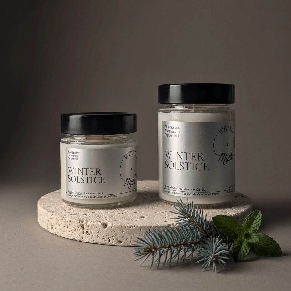 Two glass jar candles with black lids and white labels reading WINTER SOLSTICE, styled on a round stone platter with blue-green pine branches and mint leaves against a dark taupe background.