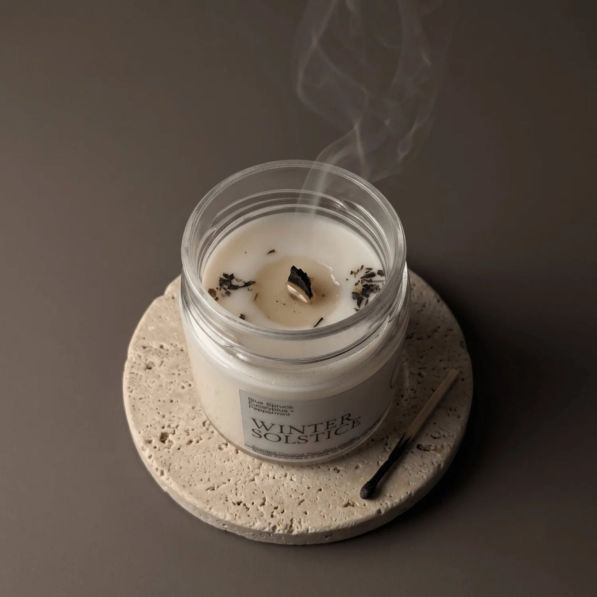 A white candle in a clear glass jar sits on a round stone coaster, with a match beside it and smoke rising from the wax. The label reads 'WINTER SOLSTICE'.