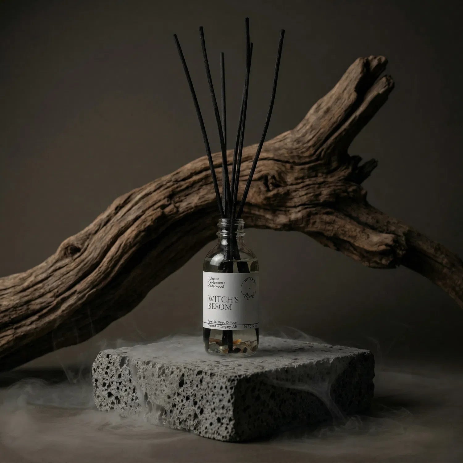 The Witch's Besom apothecary spell jar reed diffuser by Witch's Mark sitting on a stone base, enveloped in atmospheric mist with natural driftwood in the background, designed to clear stagnant energy and purify your ritual circle.