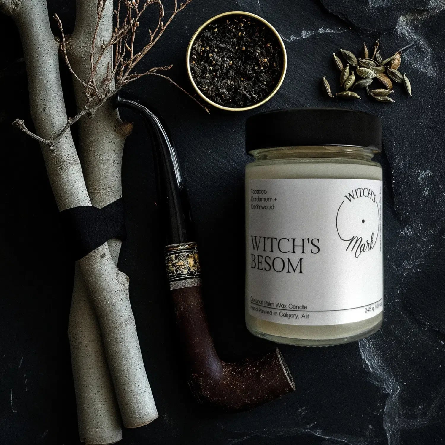 A top-down flat lay highlighting the ingredients of the Witch's Besom candle, featuring the jar surrounded by a wooden pipe with loose tobacco, green cardamom pods, and tied twigs representing a besom broom on a dark slate background.