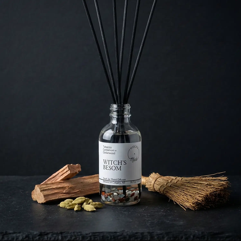 The Witch's Besom spell jar reed diffuser by Witch's Mark showcased against a dark slate background, surrounded by its core protective ingredients: a miniature sweeping besom, green cardamom pods, and pieces of raw cedarwood.