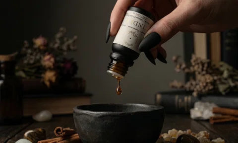 A person with manicured hands and black finger nails dispenses a single amber droplet from an essential oil bottle labelled "Cinder"