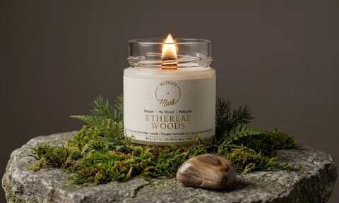 A lit candle called "Ethereal Woods" sits on top of a bed of moss and a rock. Next to the candle is a petrified wood crystal.