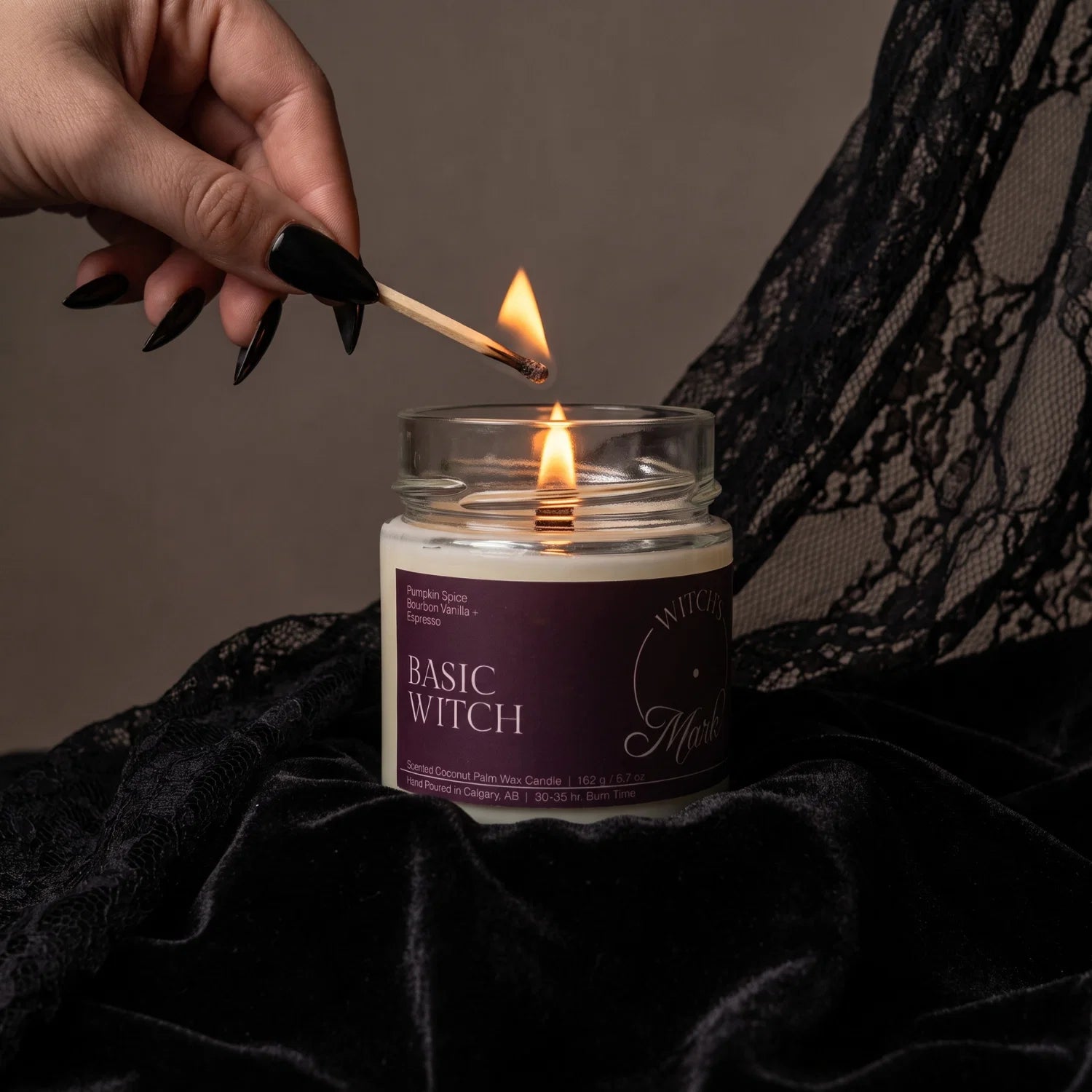 A hand with black fingernails holds a lit match to ignite a Basic Witch crystal candle.