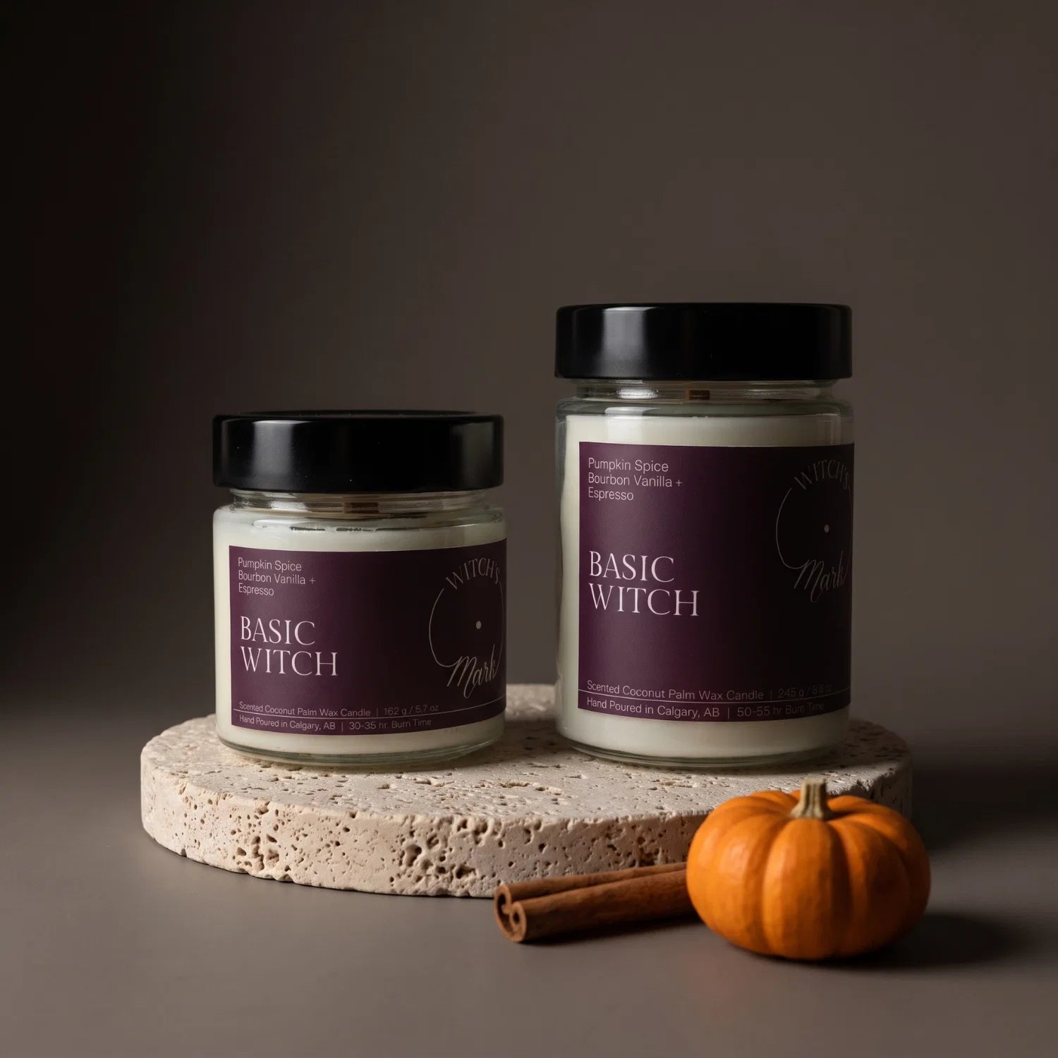 Two Basic Witch crystal candles on a porous stone coaster with a mini orange pumpkin and cinnamon stick.