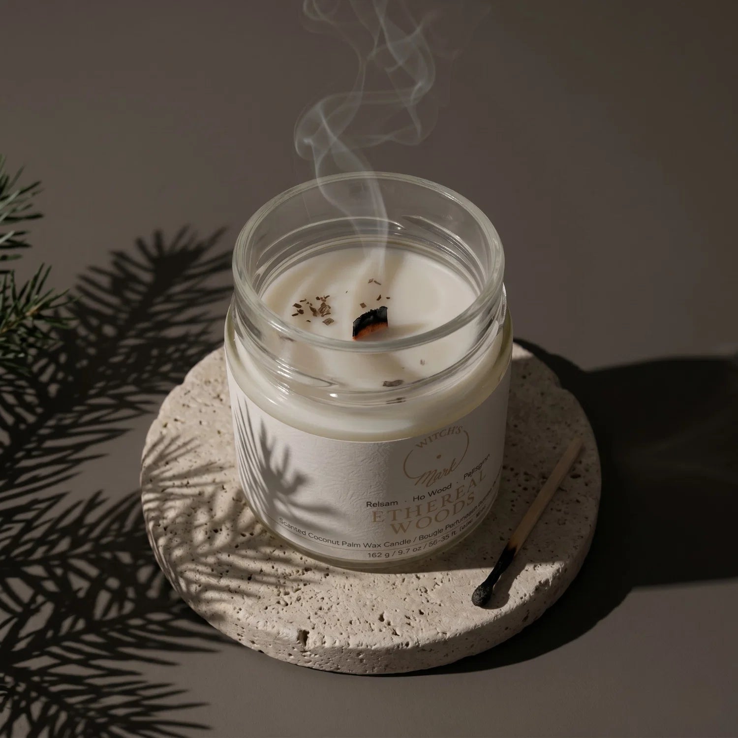 Ethereal Wood crystal candle extinguished on a porous stone coaster, surrounded by pine branches on a dark background.