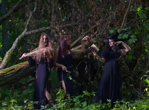Three witches in dark clothing and hats in a forest setting.