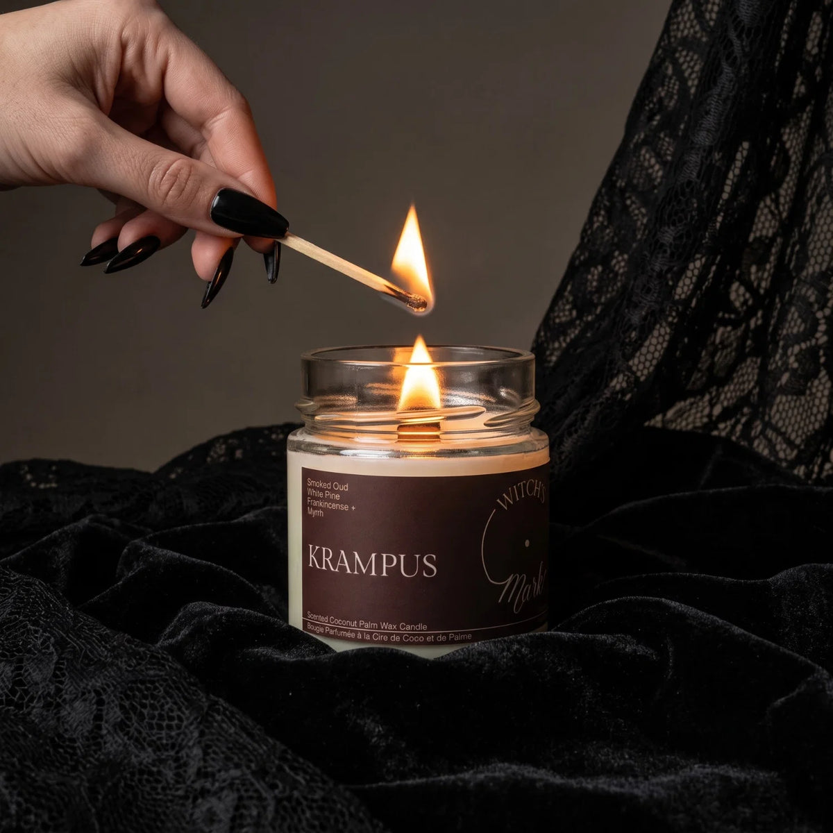 A lit Witch's Mark scented candle in a clear glass jar sits on black lace fabric, with a hand holding a match above the flame. The label reads 'KRAMPUS'.